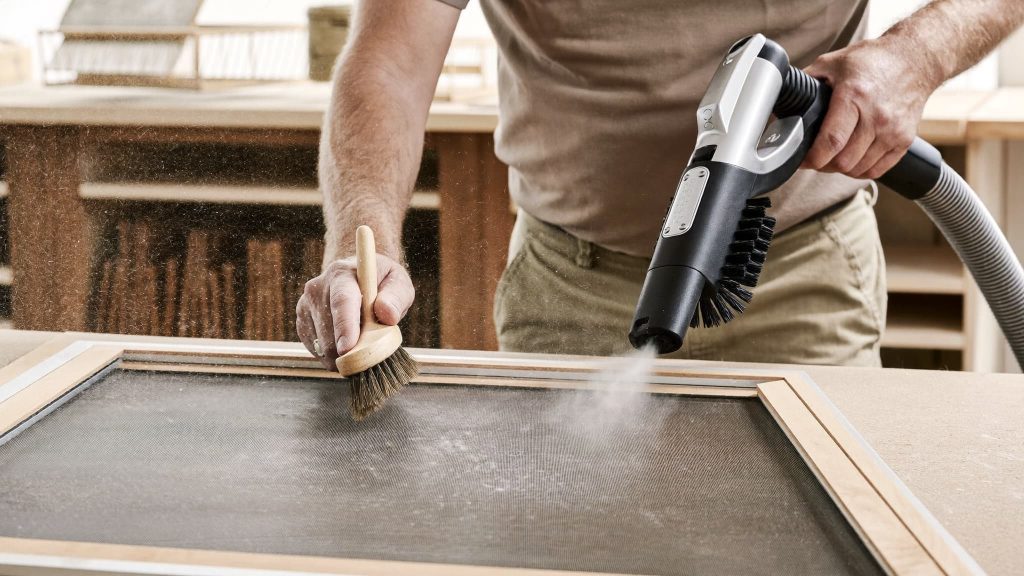 Person cleaning reusable air filter with soft brush and handheld vacuum in workshop setting.