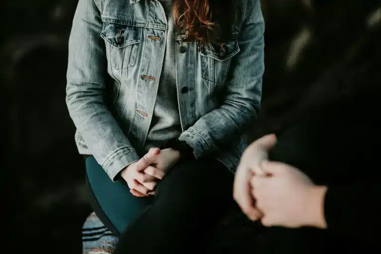 Two people seated close together, both with clasped hands, one wearing a denim jacket