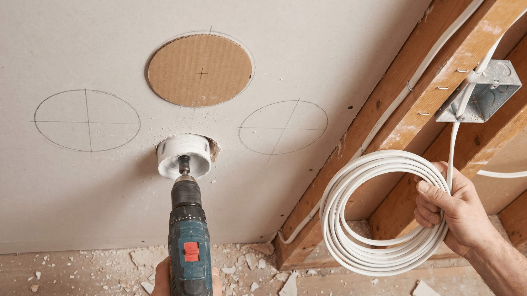 Person cutting a circular hole in the ceiling with a hole saw while holding coiled electrical wire, with ceiling markings and exposed joists visible.