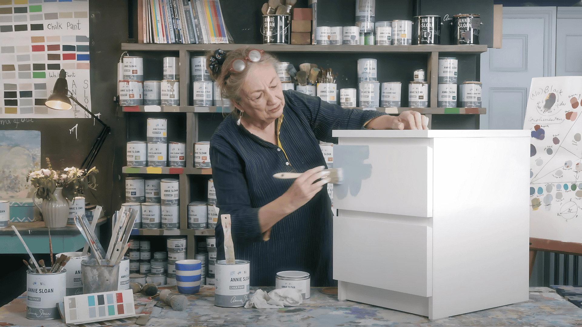 A woman in a workshop uses a paintbrush to apply a second coat of grey paint to a white two-drawer IKEA dresser.