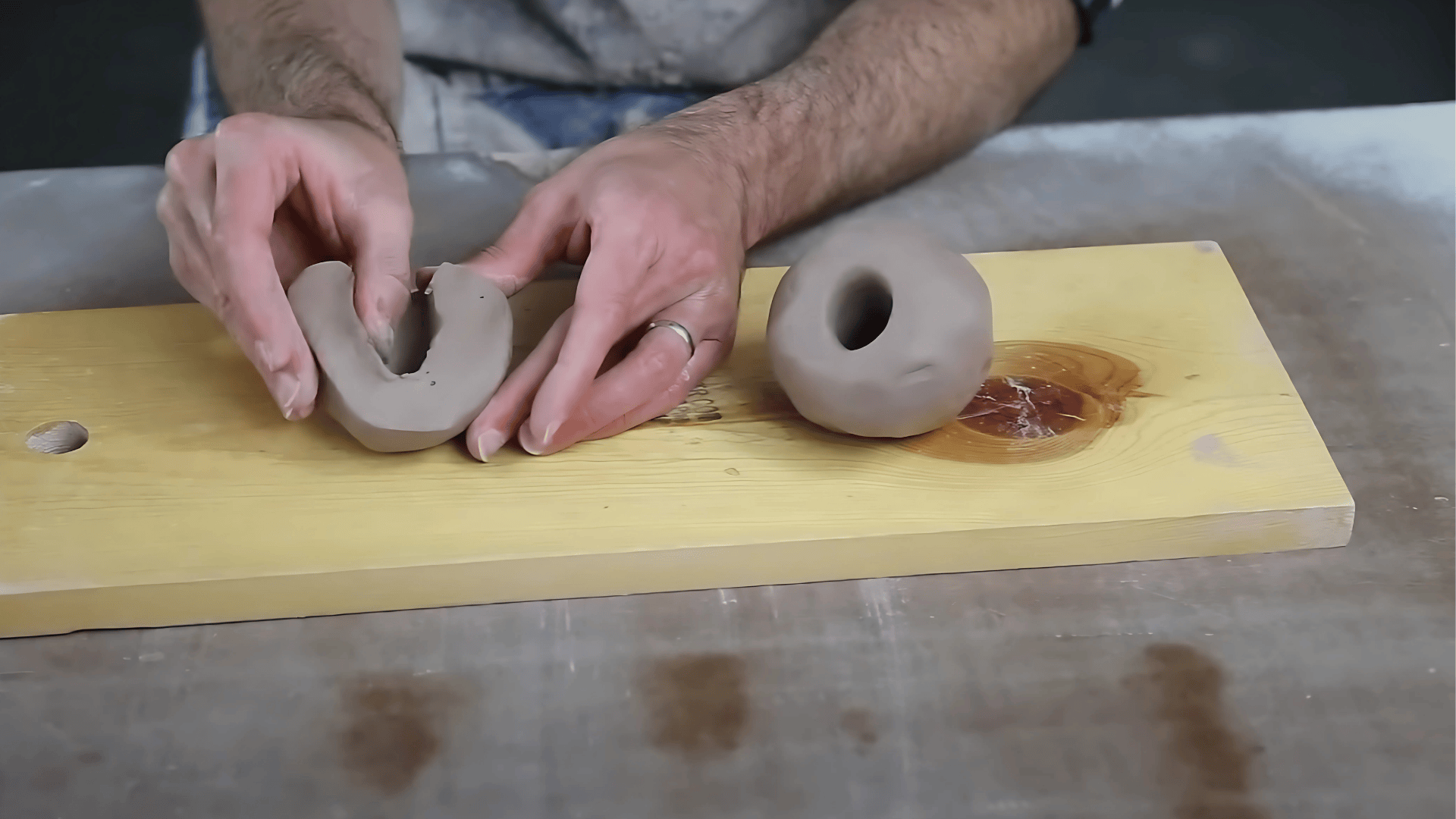A close-up shot of a person's hands shaping and pinching gray clay forms on a light-colored wooden board placed on a work surface.