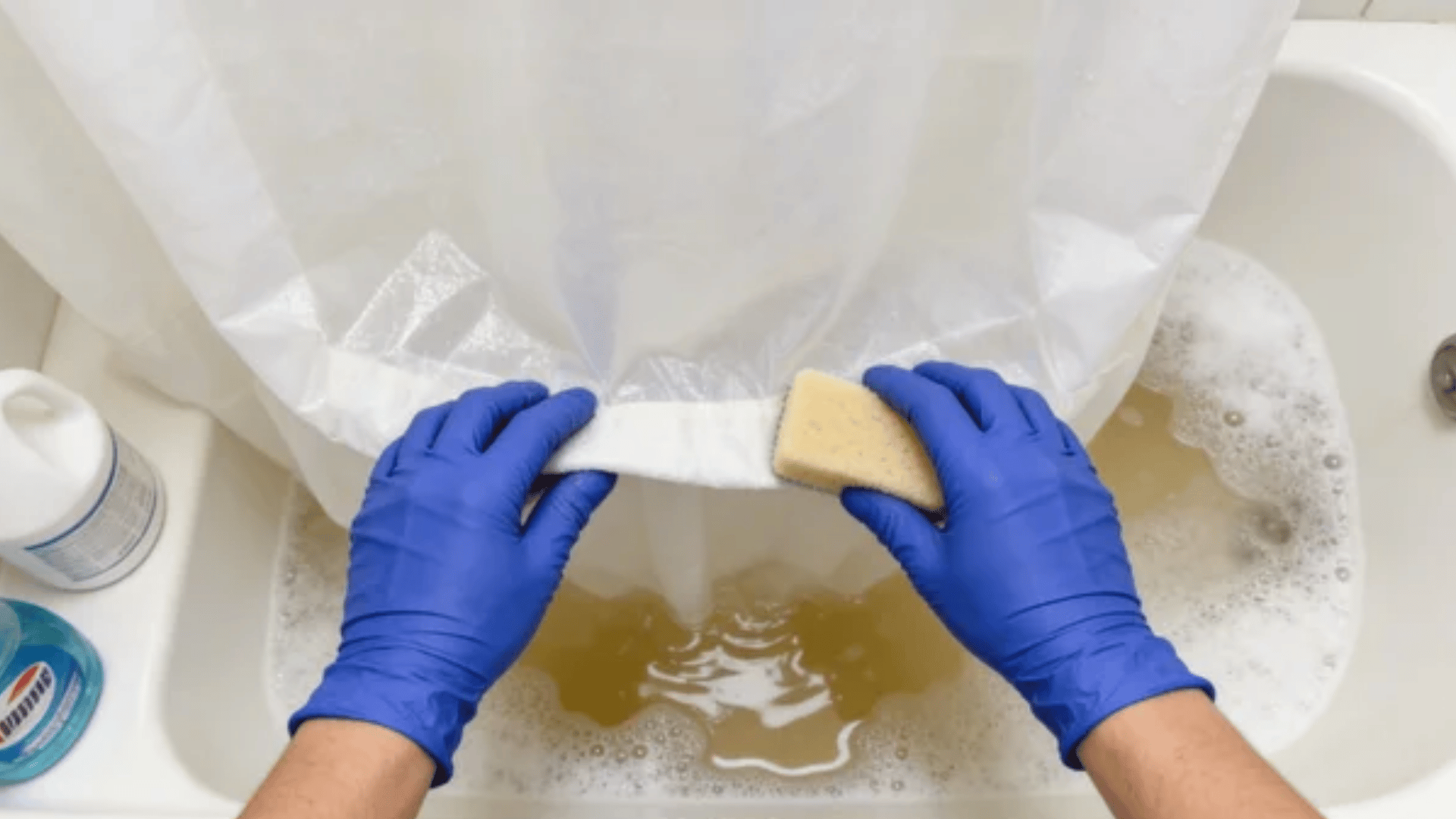 Blue gloved hands scrubbing a shower curtain in a bathtub with soapy water