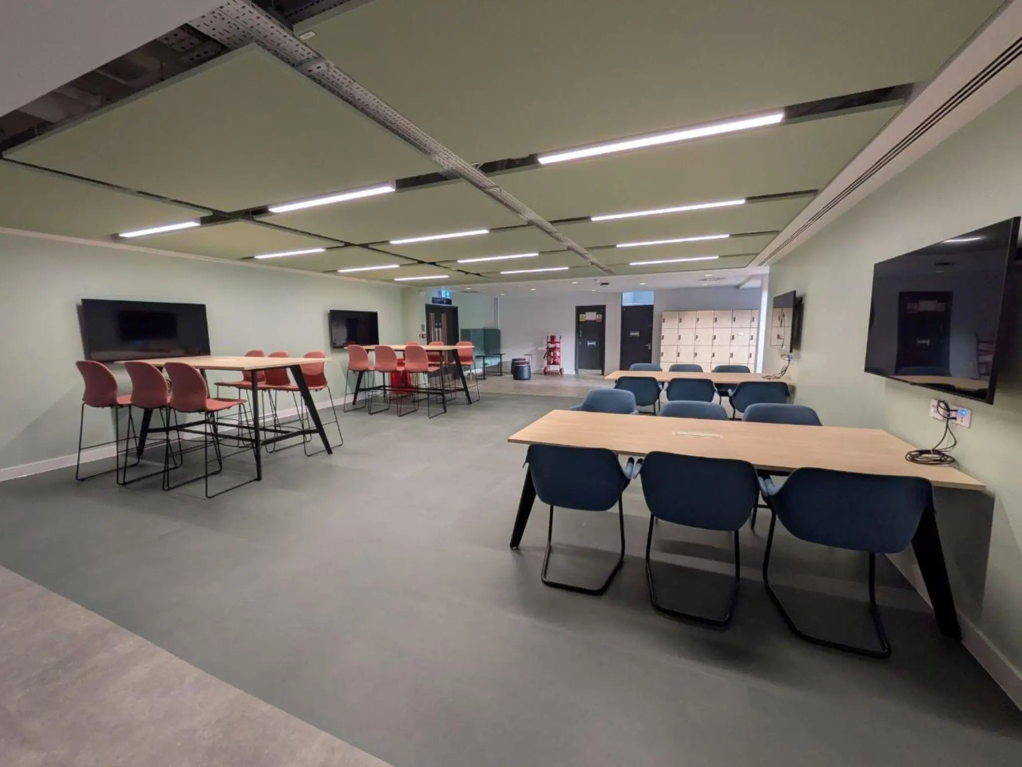 Modern office breakout area with high tables, red chairs, and wall-mounted screens
