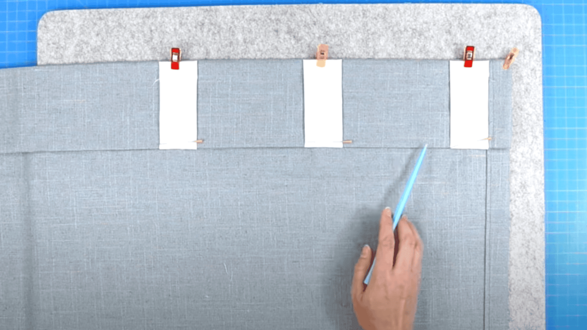 Aligning white fabric tabs on a grey curtain panel with a light blue marker and red sewing clips.