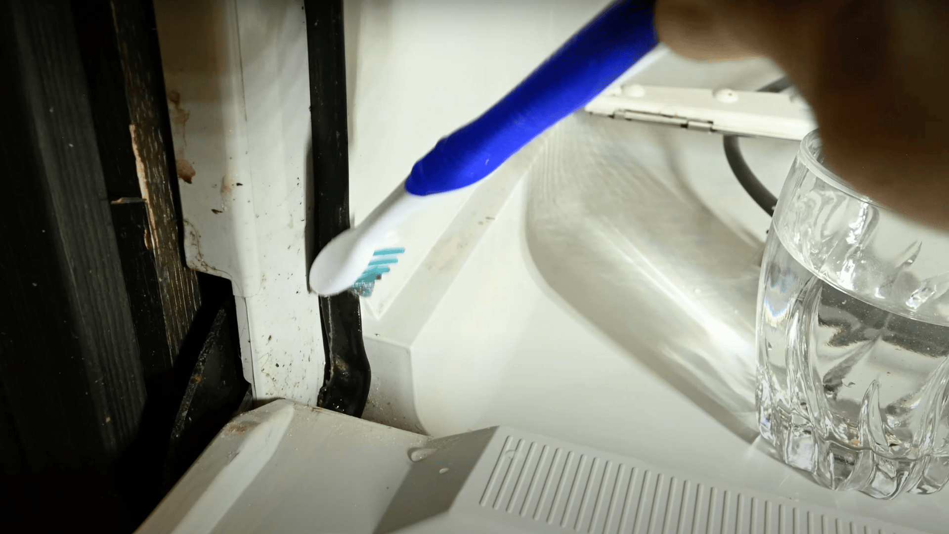 An image showing a person using a blue toothbrush to scrub grime and buildup along the black rubber door seal of an open dishwasher.