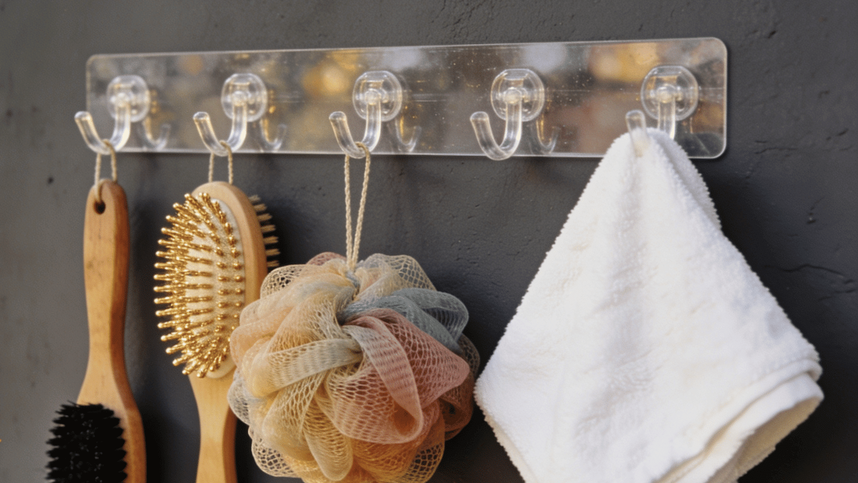 Clear adhesive wall hook strip holding brushes, a bath pouf, and a white towel.