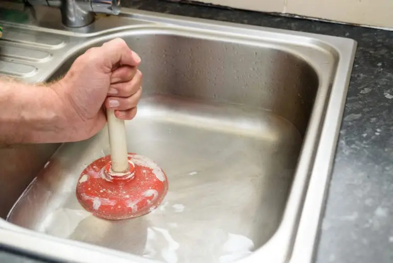 Hand using red plunger in water-filled stainless steel sink in kitchen