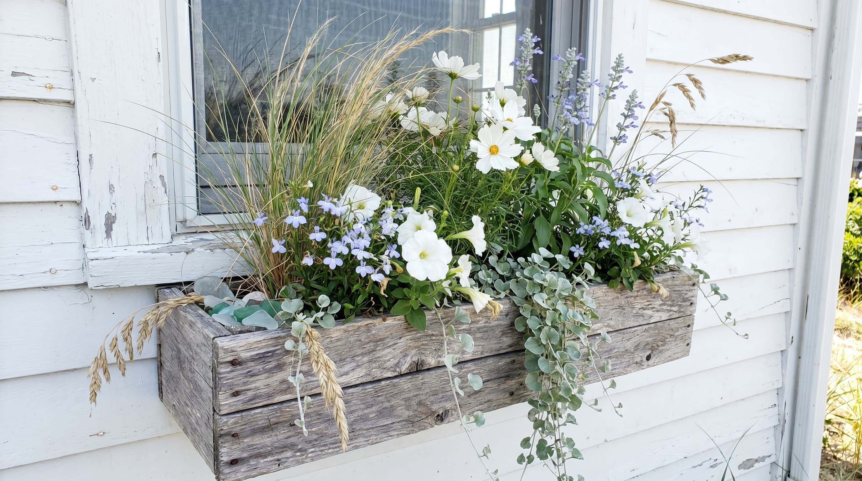 A rustic wooden coastal-inspired window box filled with white and pale purple flowers, ornamental grasses, and cascading greenery against a white house exterior.
