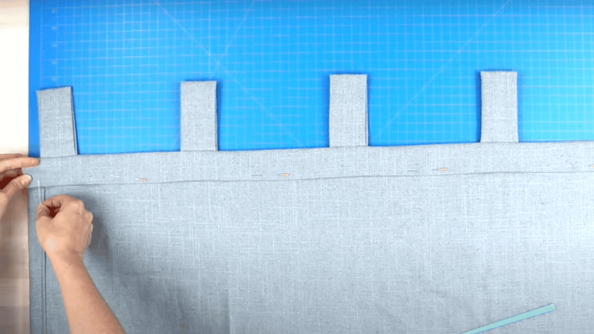 An image of a person's hands sewing the top edge of a light grey curtain panel with white fabric tabs on a blue cutting mat.