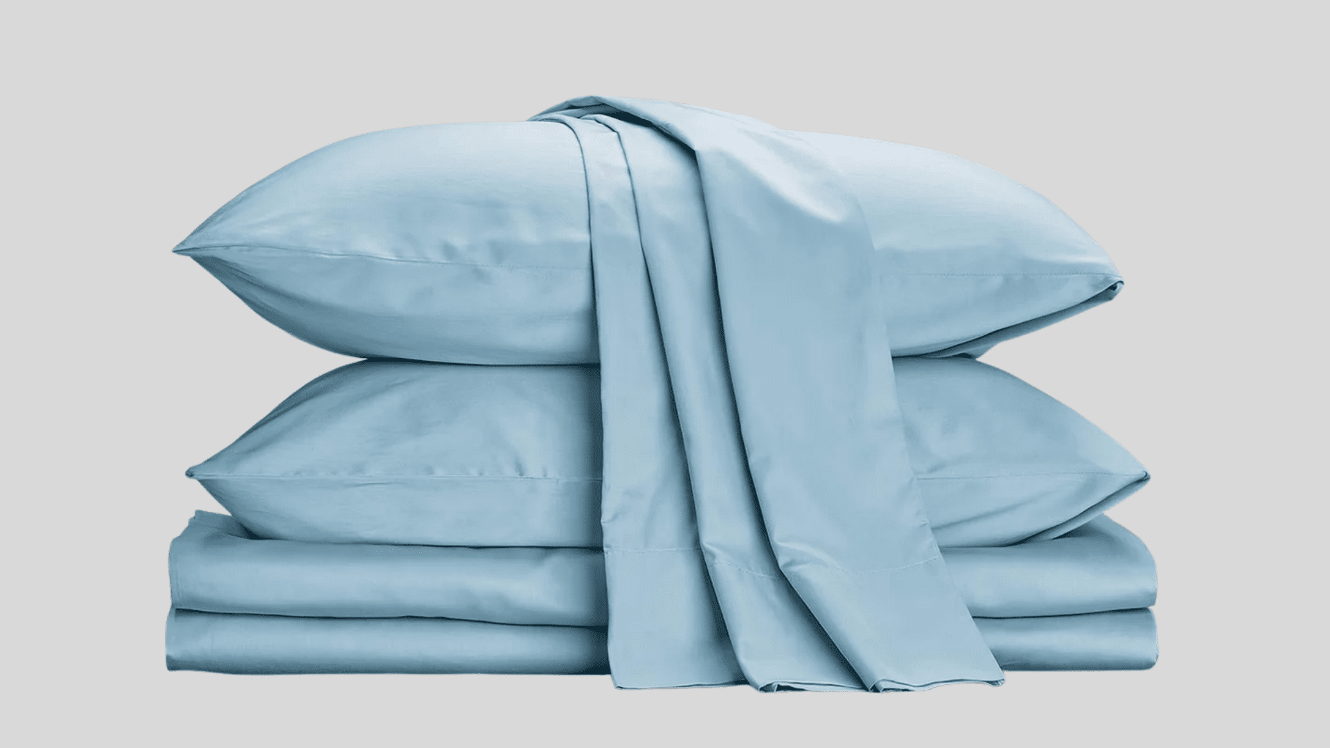 Neatly folded stack of dusty blue Cotton sheets, including two pillowcases and a fitted sheet, against a light gray background.