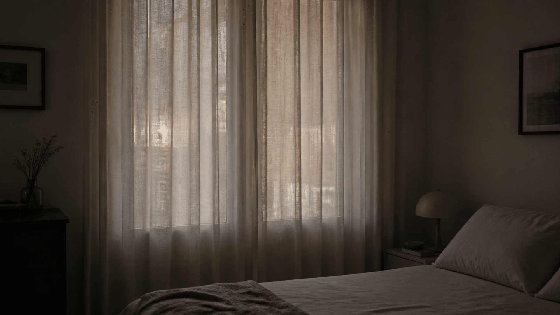Softly lit bedroom with sheer curtains covering window, bed with neutral bedding and bedside lamp in muted tones.