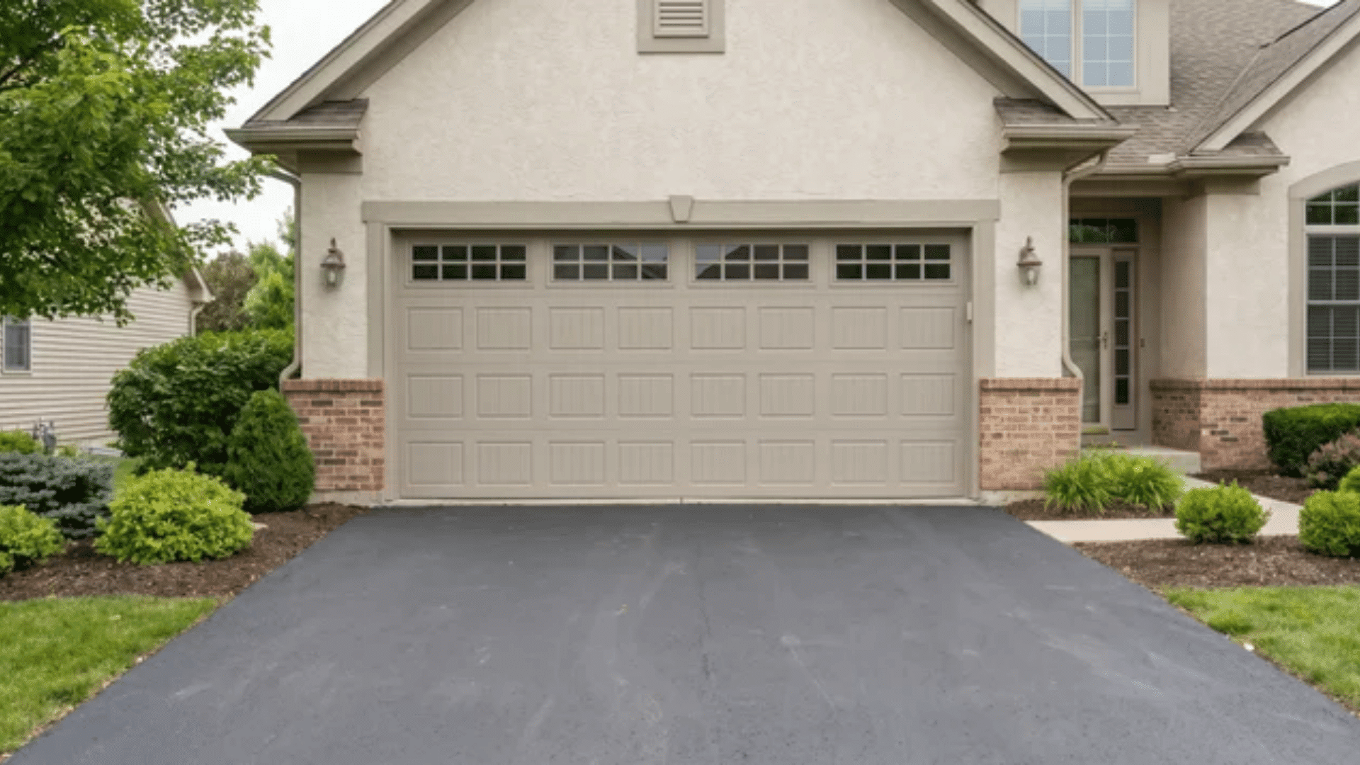 Closed beige two-car garage door on a suburban home with brick accents, wall lanterns, and landscaped front yard.