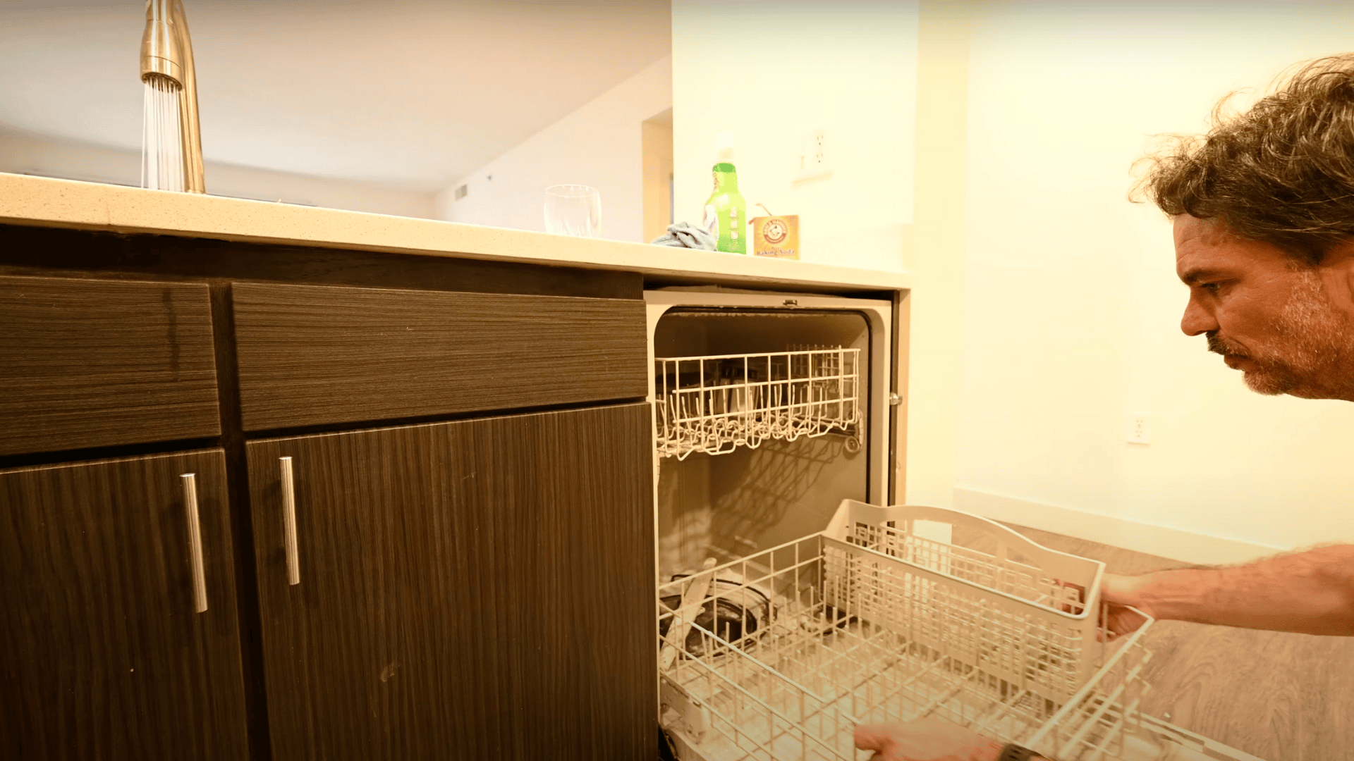 An image of a man bending down to slide the top rack back into an open dishwasher next to dark wood kitchen cabinets.