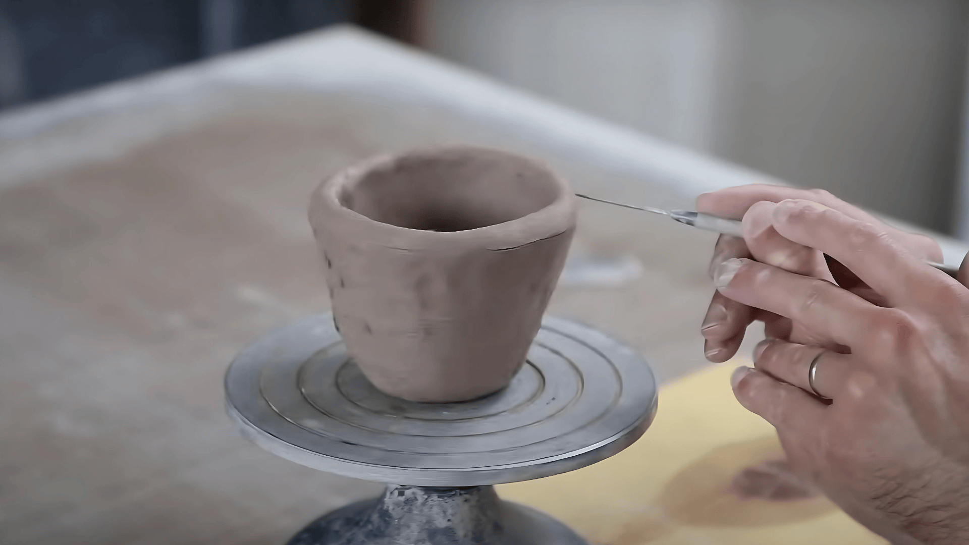 A close-up shot of a person's hands using a needle tool to trim the rim of a handmade gray clay pot on a silver banding wheel.