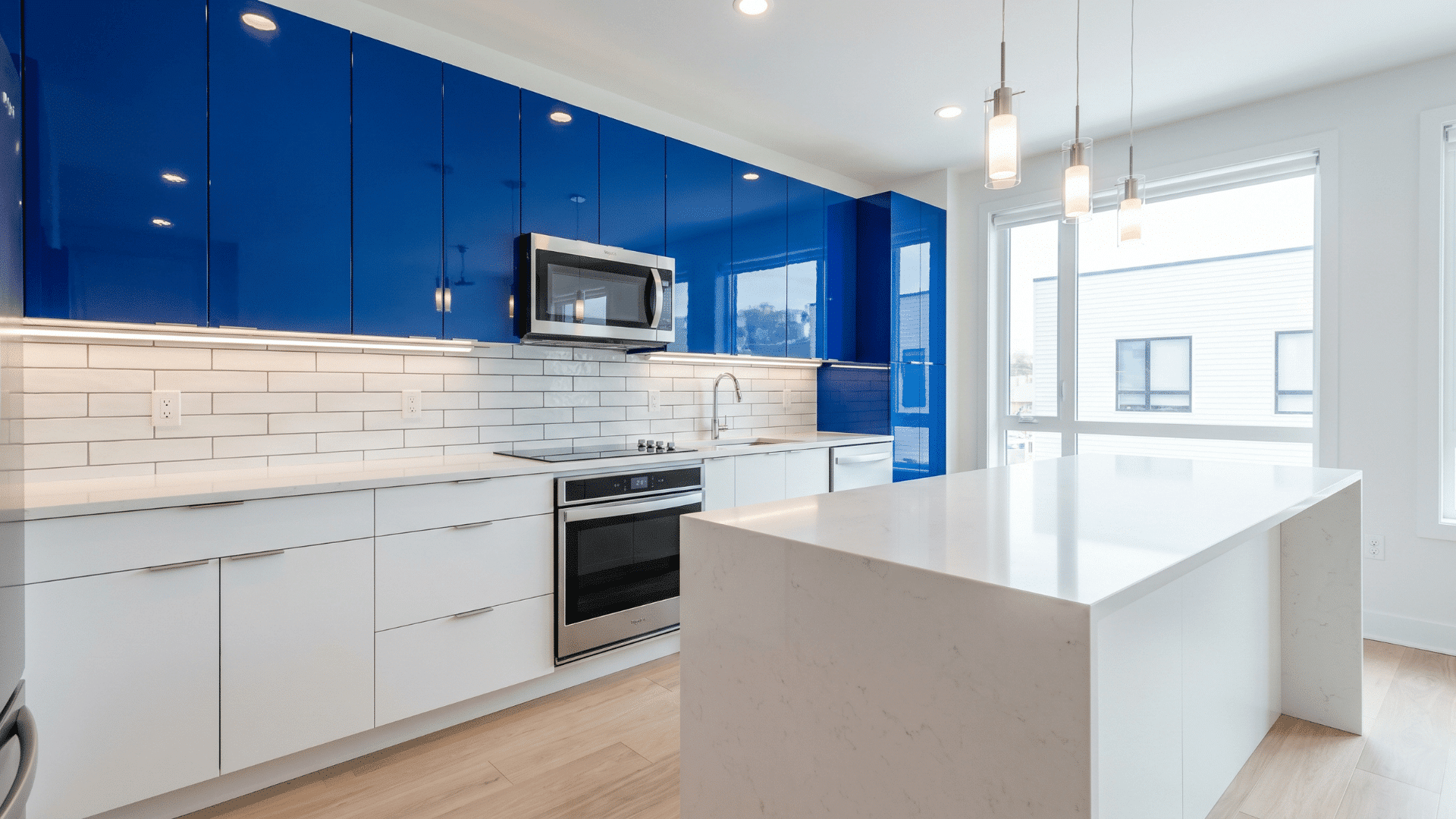 Glossy blue upper cabinets paired with white lower cabinets in a modern kitchen, featuring a large white island, stainless steel appliances, and natural light from large windows.