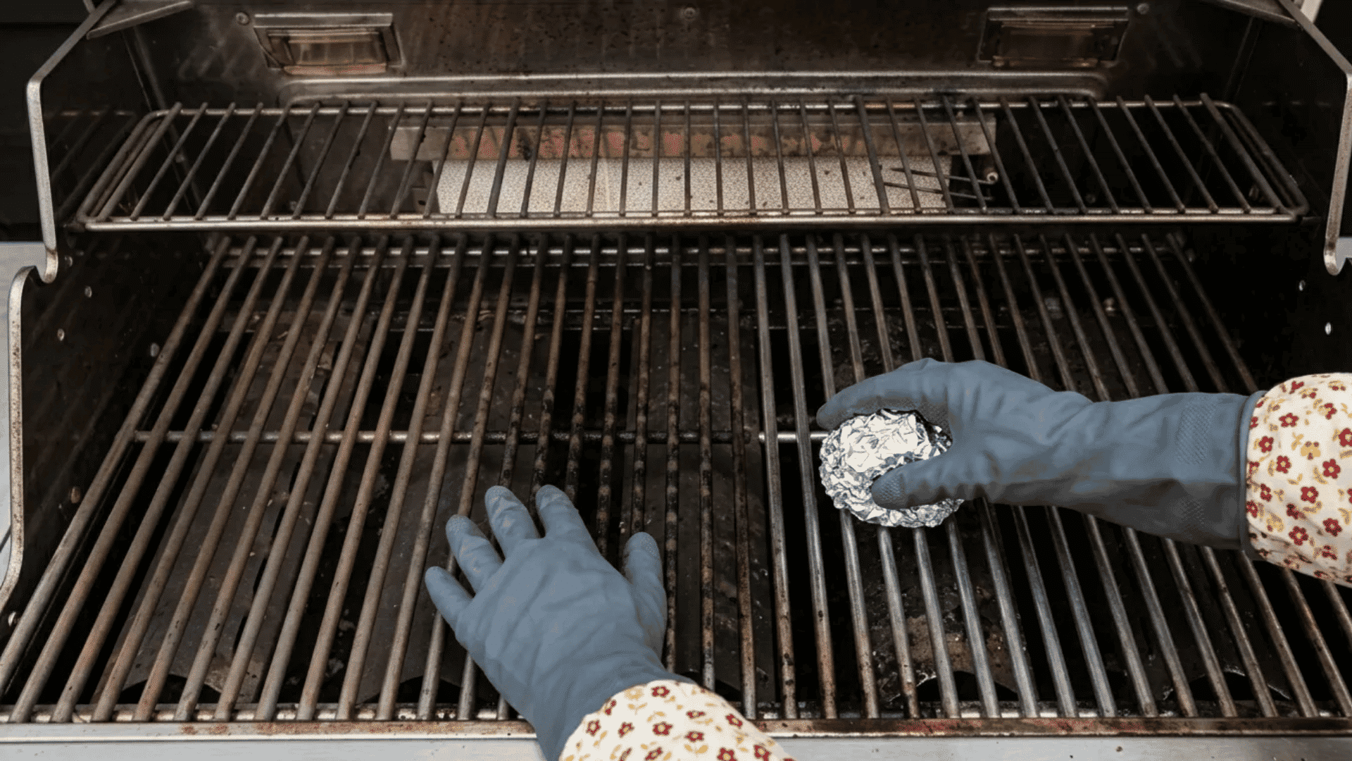 Person scrubbing dirty grill grates with a crumpled aluminum foil ball while wearing protective gloves.