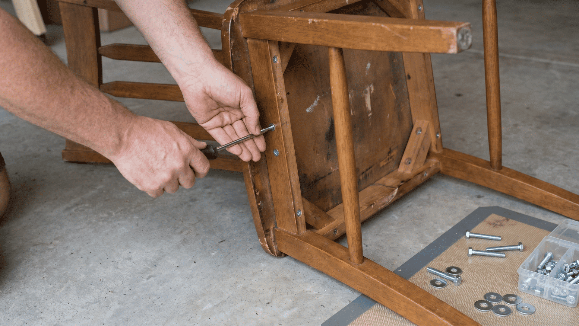 Hands tightening screws under an upside-down chair with repair tools nearby