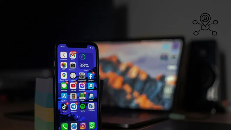 Smartphone displaying various app icons on screen in front of blurred laptop in dimly lit room