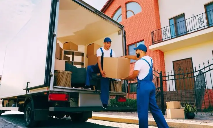 Movers in blue uniforms unloading boxes from truck in front of brick house