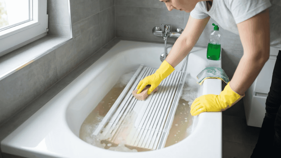 Person cleaning blinds in a bathtub with soapy water using a sponge and yellow gloves for deep cleaning.