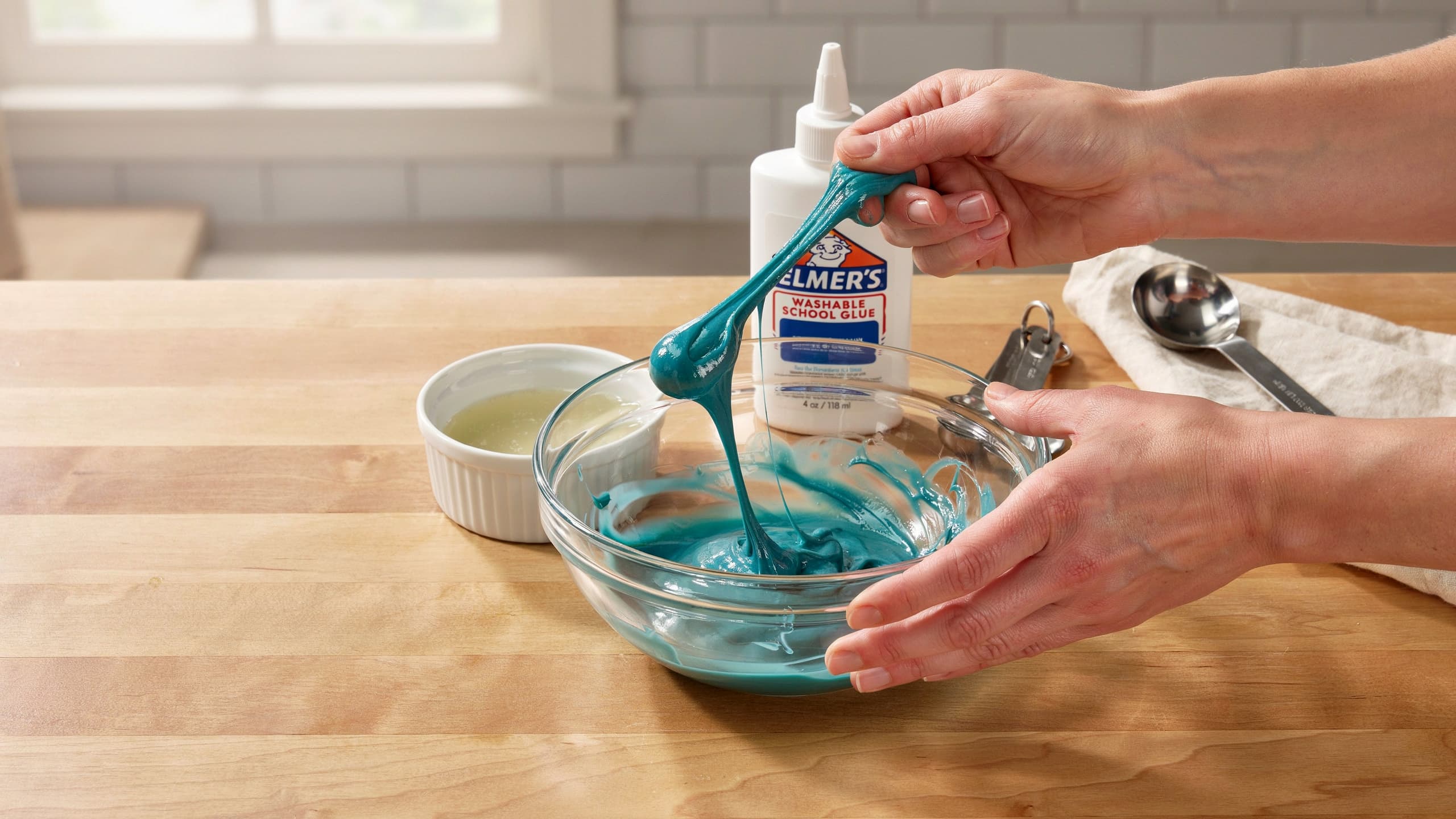 Hands mixing blue slime with a spoon in a clear glass bowl on a wooden countertop, with a bottle of Elmer's school glue held above it.