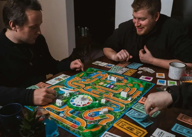 Three people playing board game at wooden table with colorful game pieces and fake money