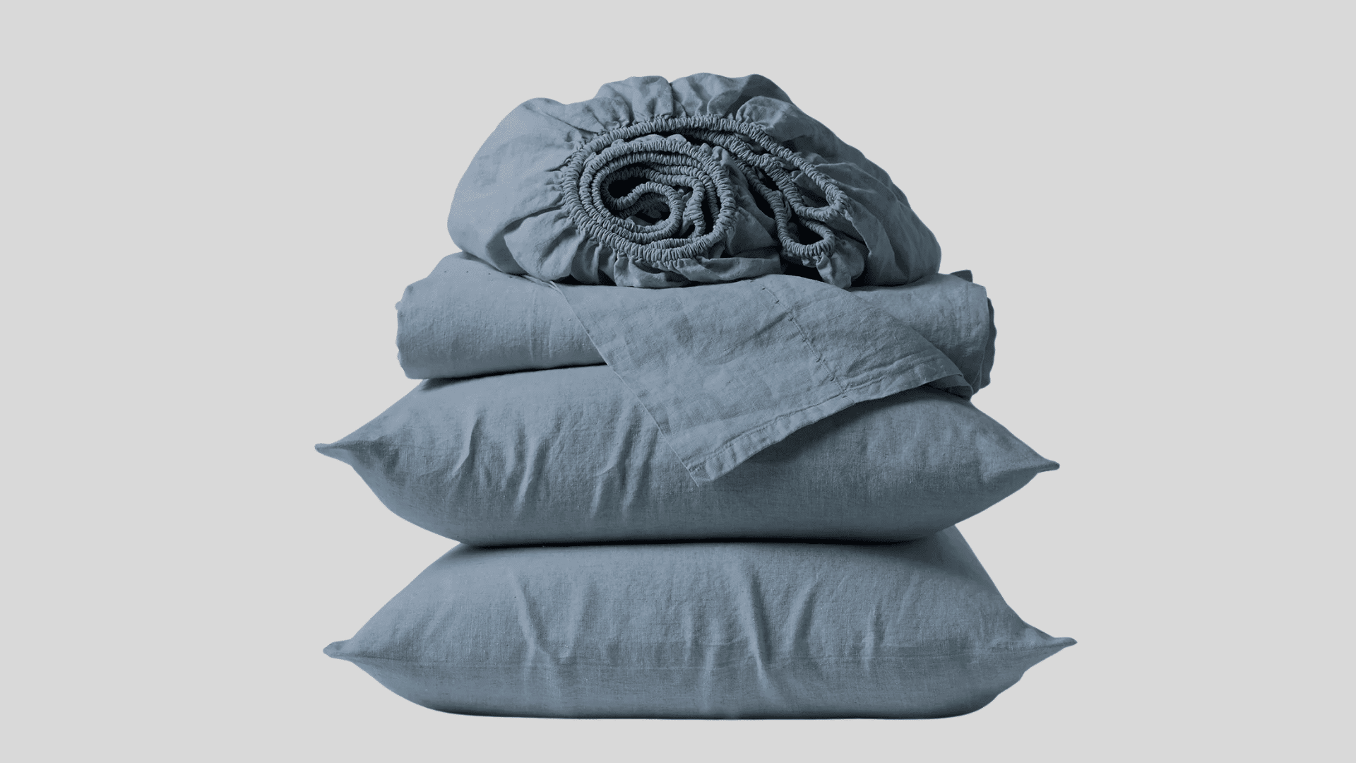 Neatly folded stack of dusty blue linen bedding, including two pillowcases and a fitted sheet, against a light gray background.