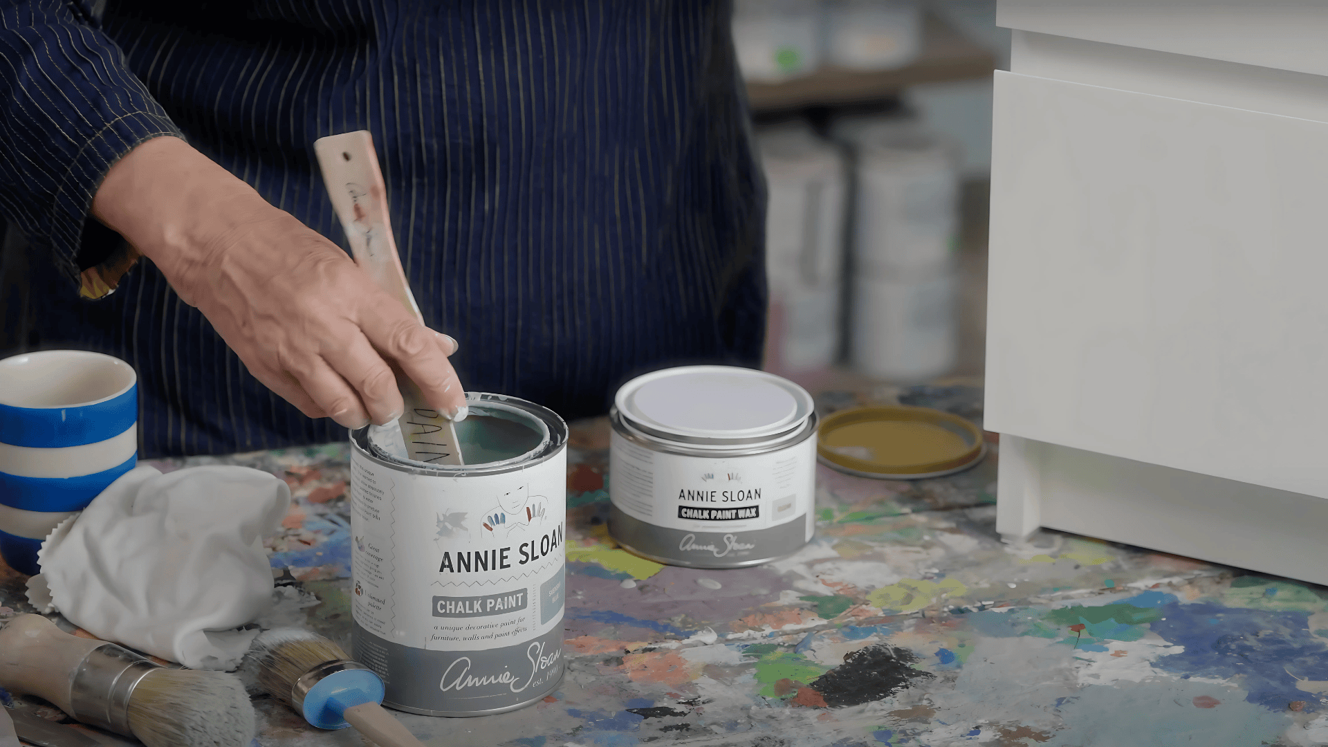 A person's hand mixes chalk paint in a tin container with a wooden stick on a workspace with a white furniture piece.
