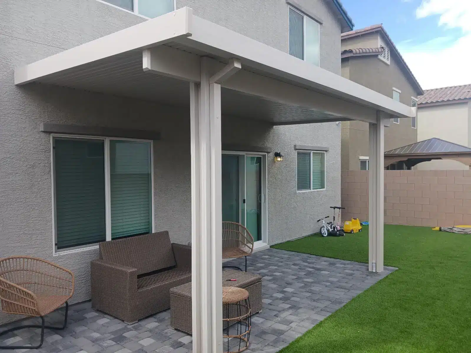 Patio with wicker furniture under white pergola on gray brick pavers in backyard setting