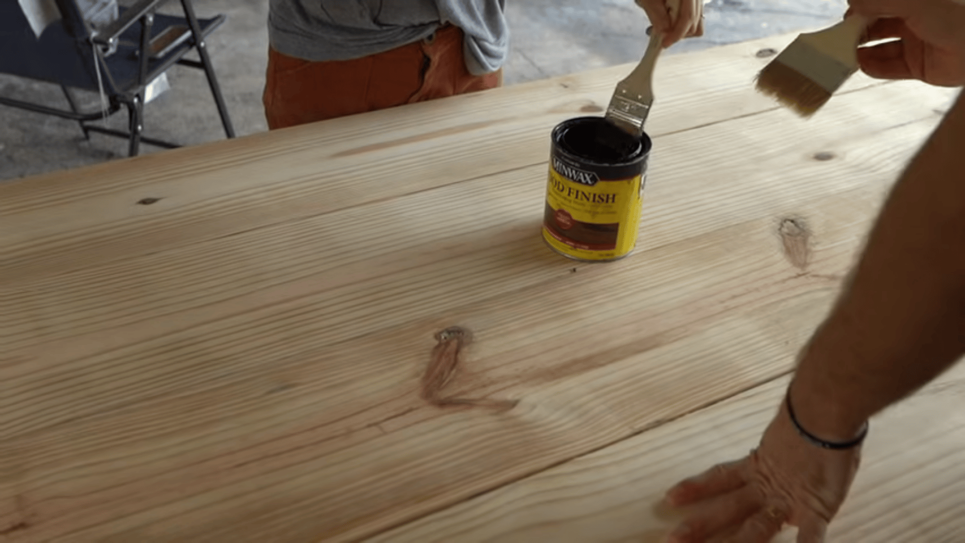 People using paintbrushes to apply wood stain from a Minwax can onto a light wooden tabletop