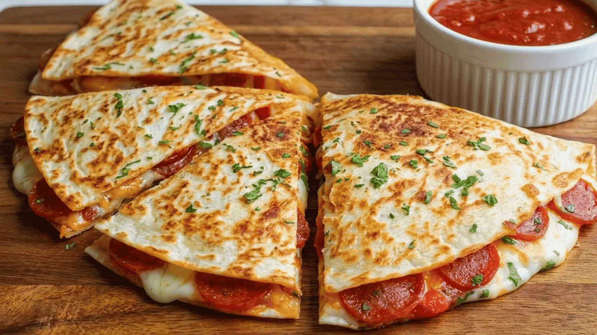 Golden pizza quesadillas sliced into triangles on a wooden board with melted cheese, pepperoni, herbs, and marinara dipping sauce on the side.