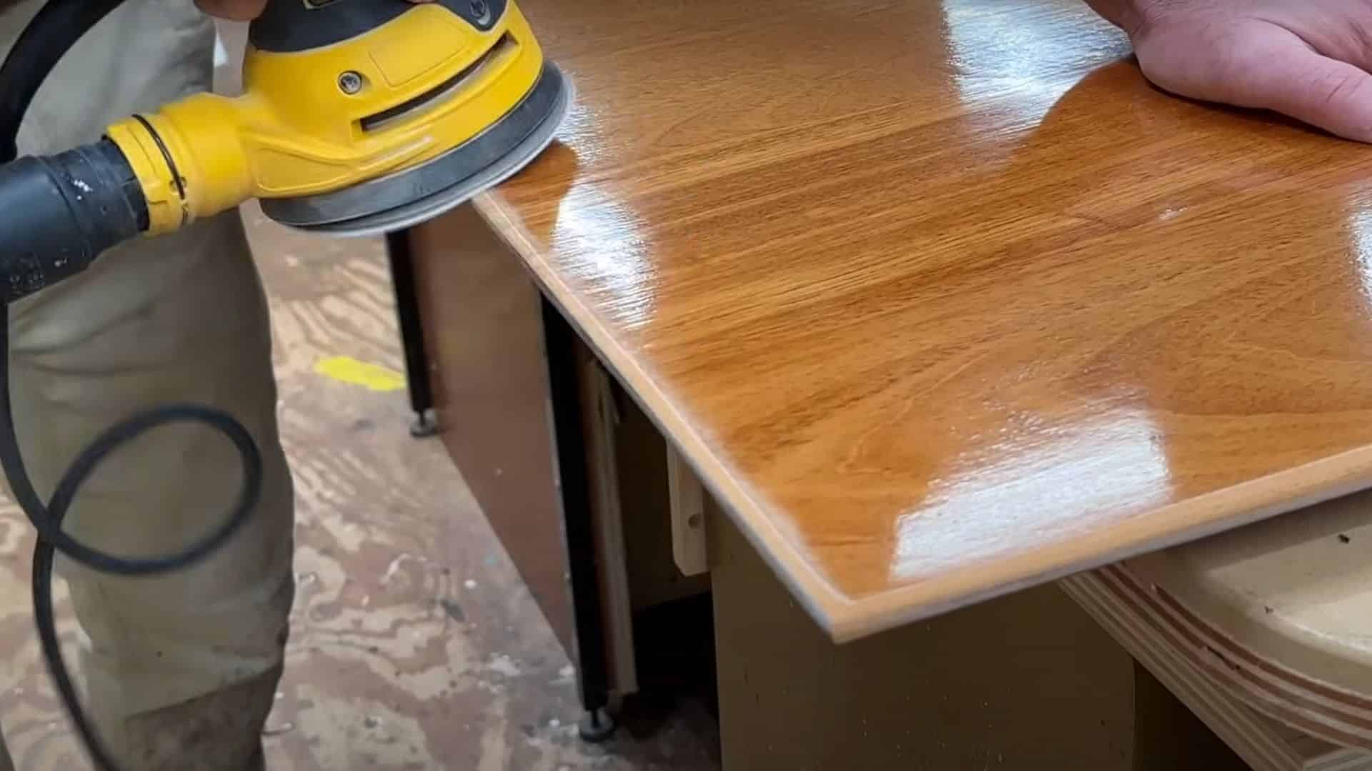 Hand using a yellow orbital sander to smooth the rounded corners and edges of a wooden tabletop.