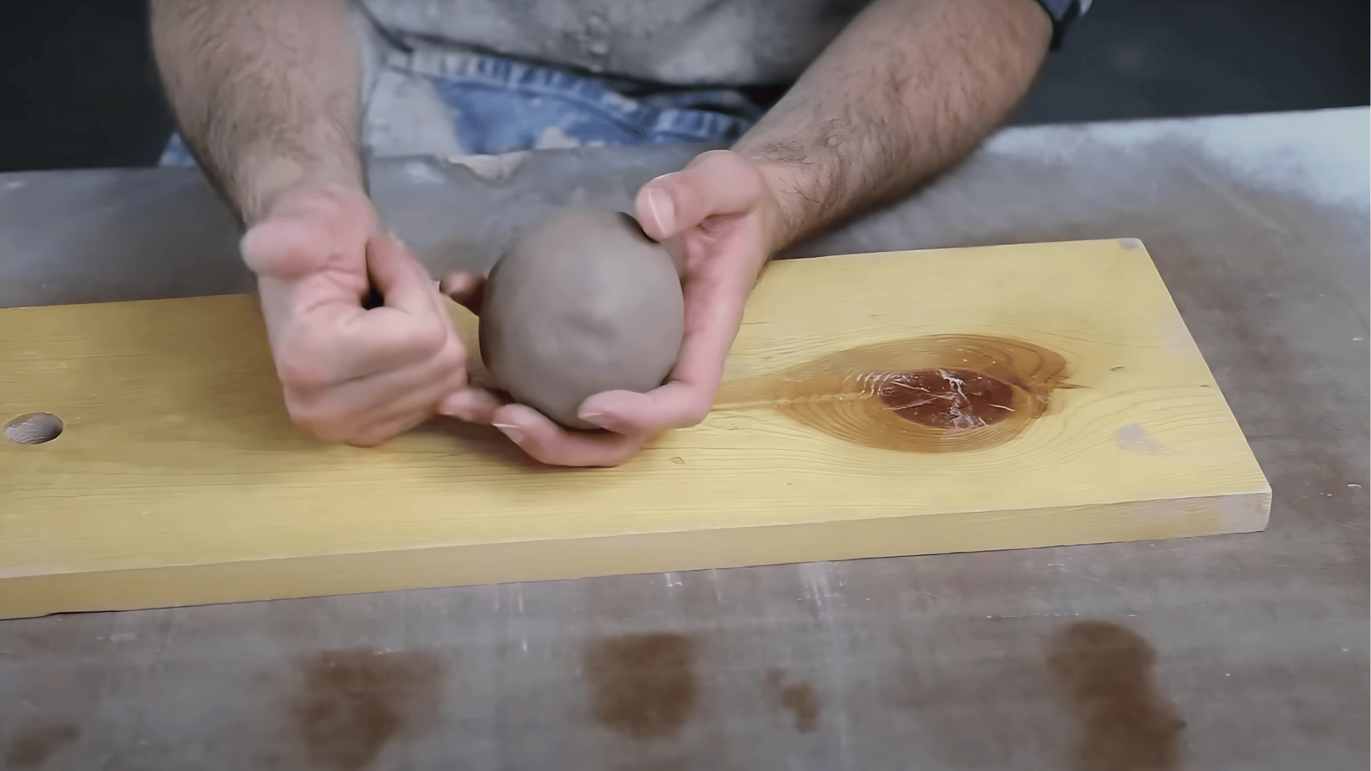 A close-up shot of a person's hands shaping a gray ball of clay on a light-colored wooden board placed on a work surface.