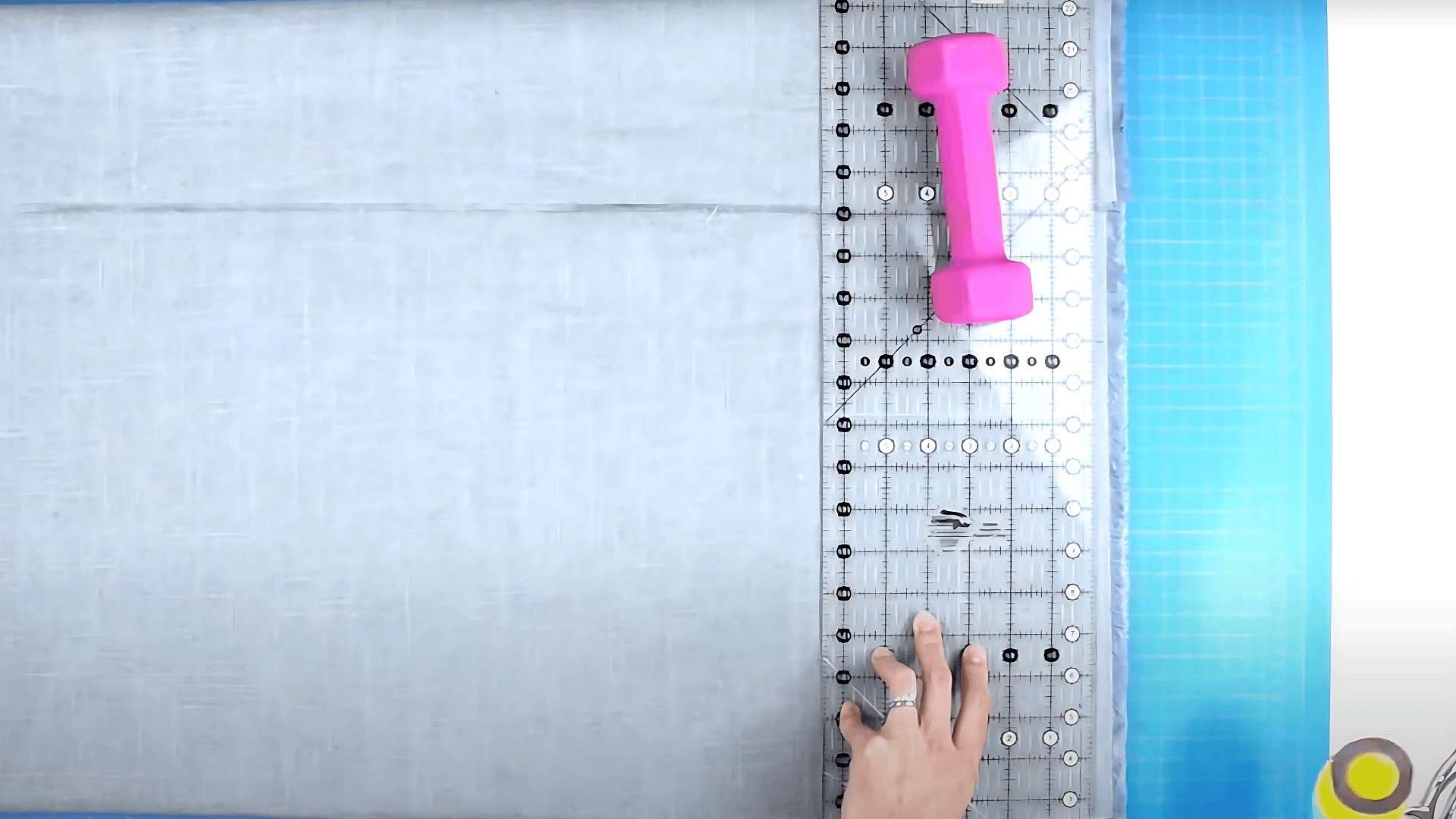 An image of a person using a clear ruler, a pink weight, and a blue cutting mat to measure and cut light grey fabric for a curtain.