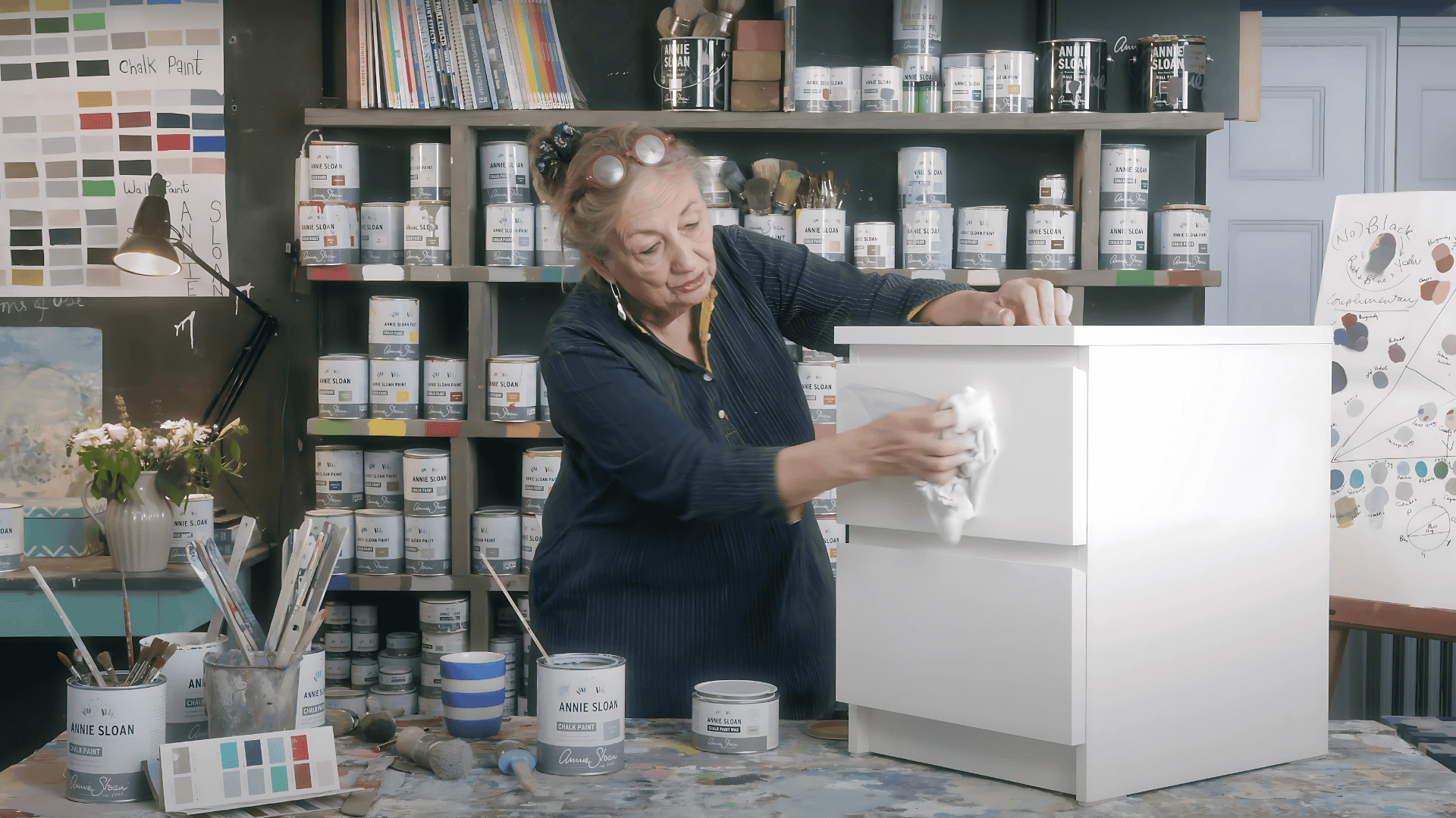 A woman in a workshop wipes down a small white two-drawer dresser with a cloth to prepare its surface for painting.