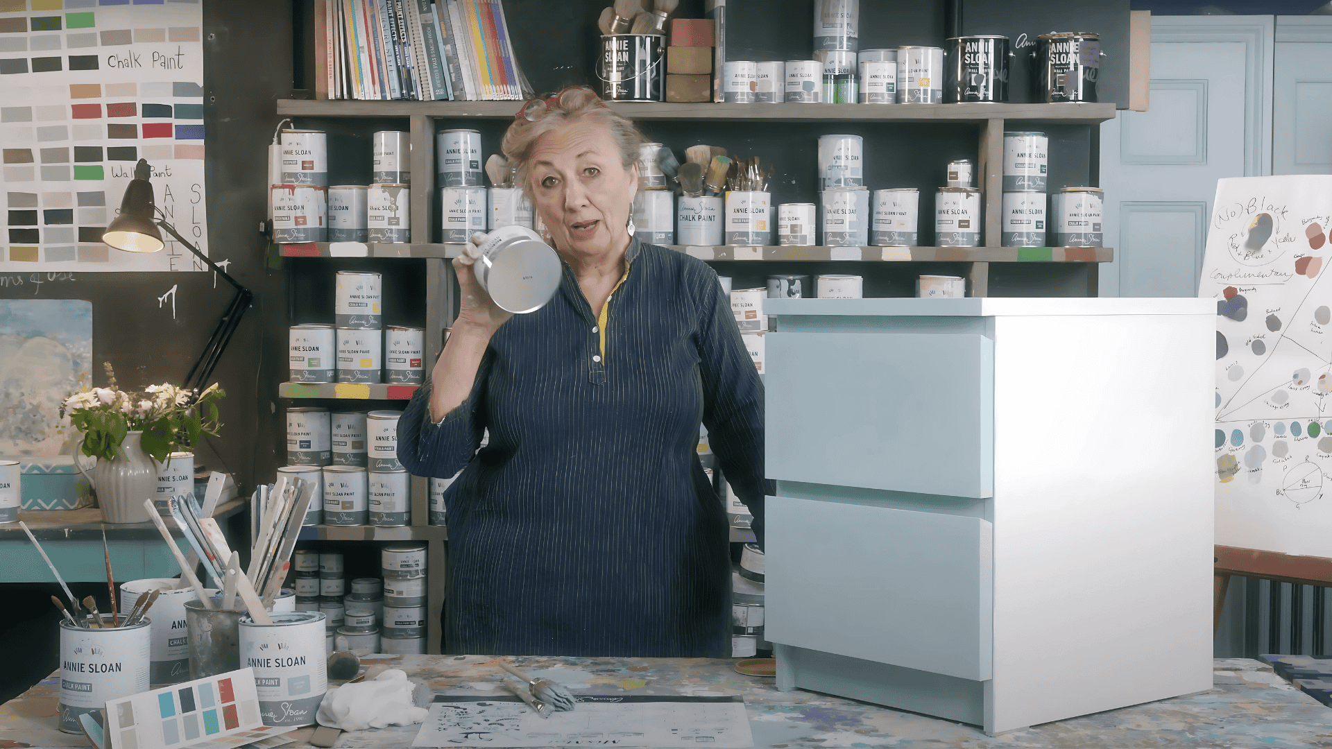 A woman in a workshop stands next to a painted two-drawer IKEA dresser, holding up a small tin of clear protective wax.