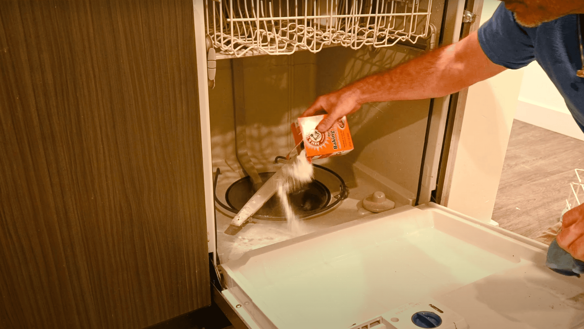 An image of a person sprinkling a cup of baking soda from an orange box onto the bottom floor of an empty, open dishwasher.
