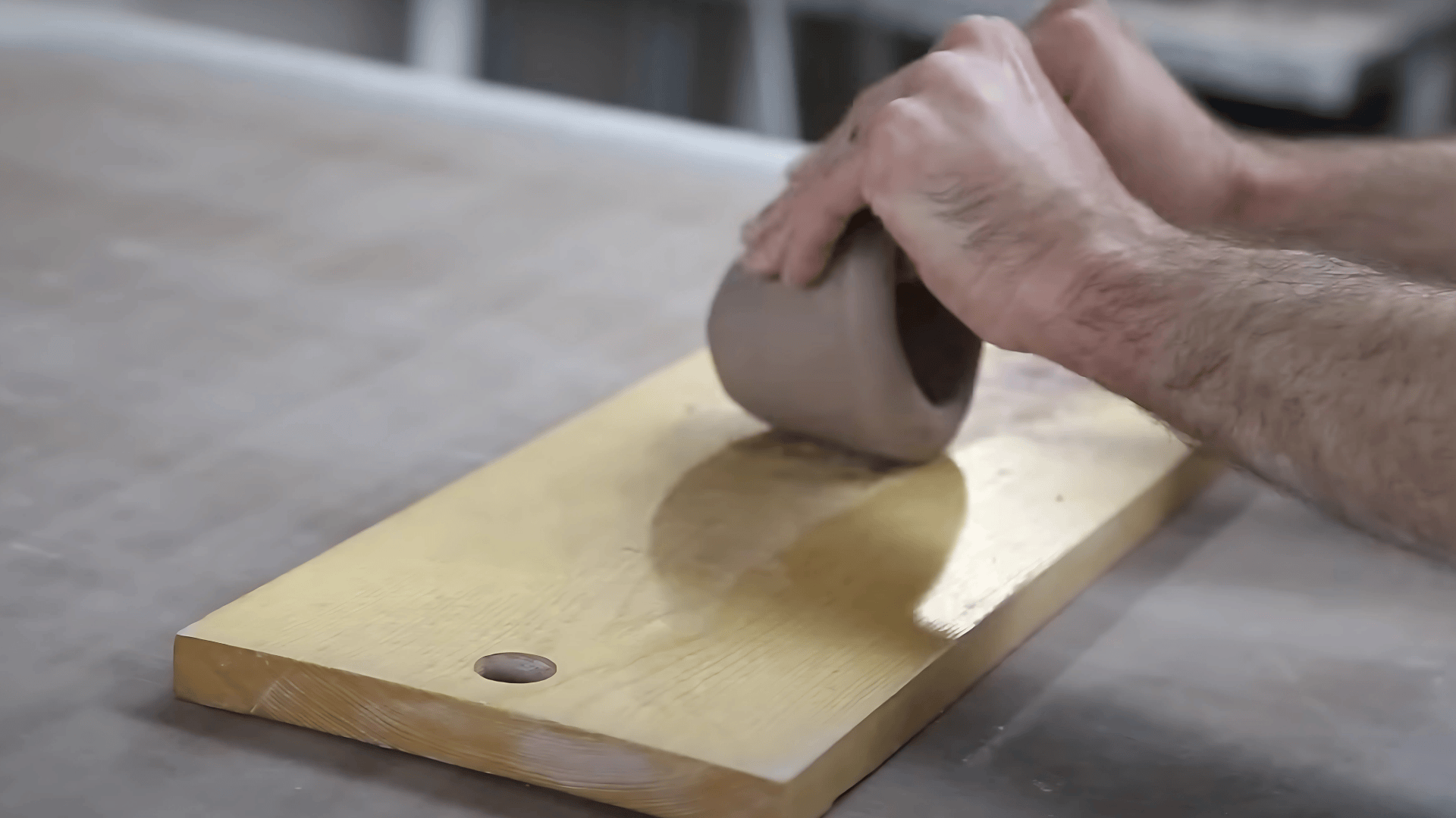 A close-up shot of a person's hands shaping and pinching gray clay forms on a light-colored wooden board placed on a work surface.