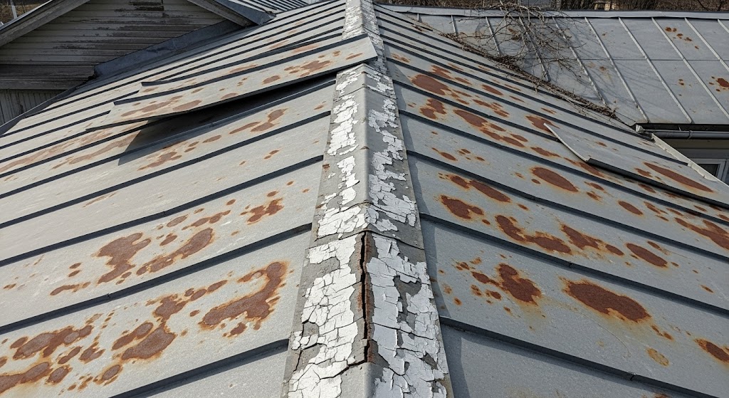 A close-up shot of an old, weathered corrugated metal roof showing severe rust spots, peeling white paint, and corroded seams.