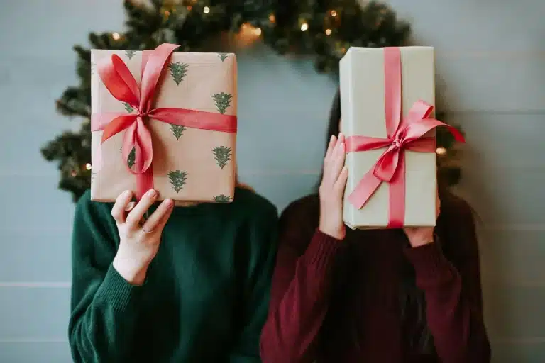 Two people holding wrapped gifts with red bows in cozy holiday setting