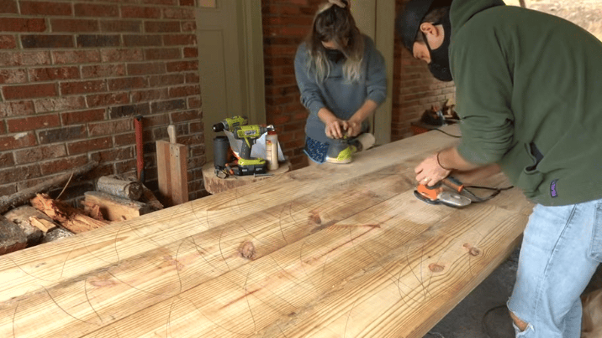 Two people using electric orbital sanders to smooth the surface of a long wooden tabletop.