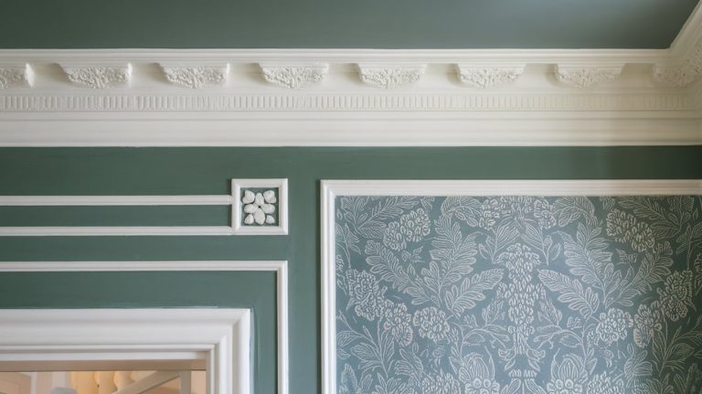 A close-up shot of a sage green wall featuring white crown molding and trim framing blue floral wallpaper.