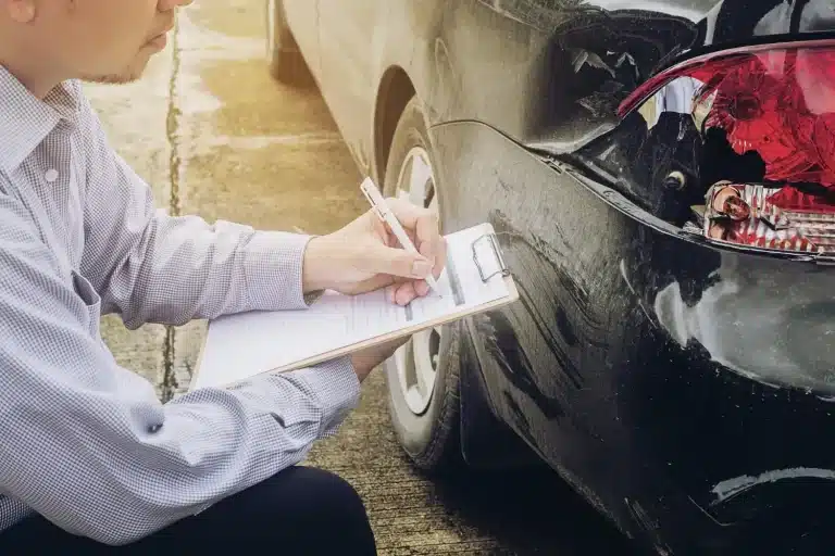 Man inspecting and writing on clipboard next to car with rear-end damage outdoors