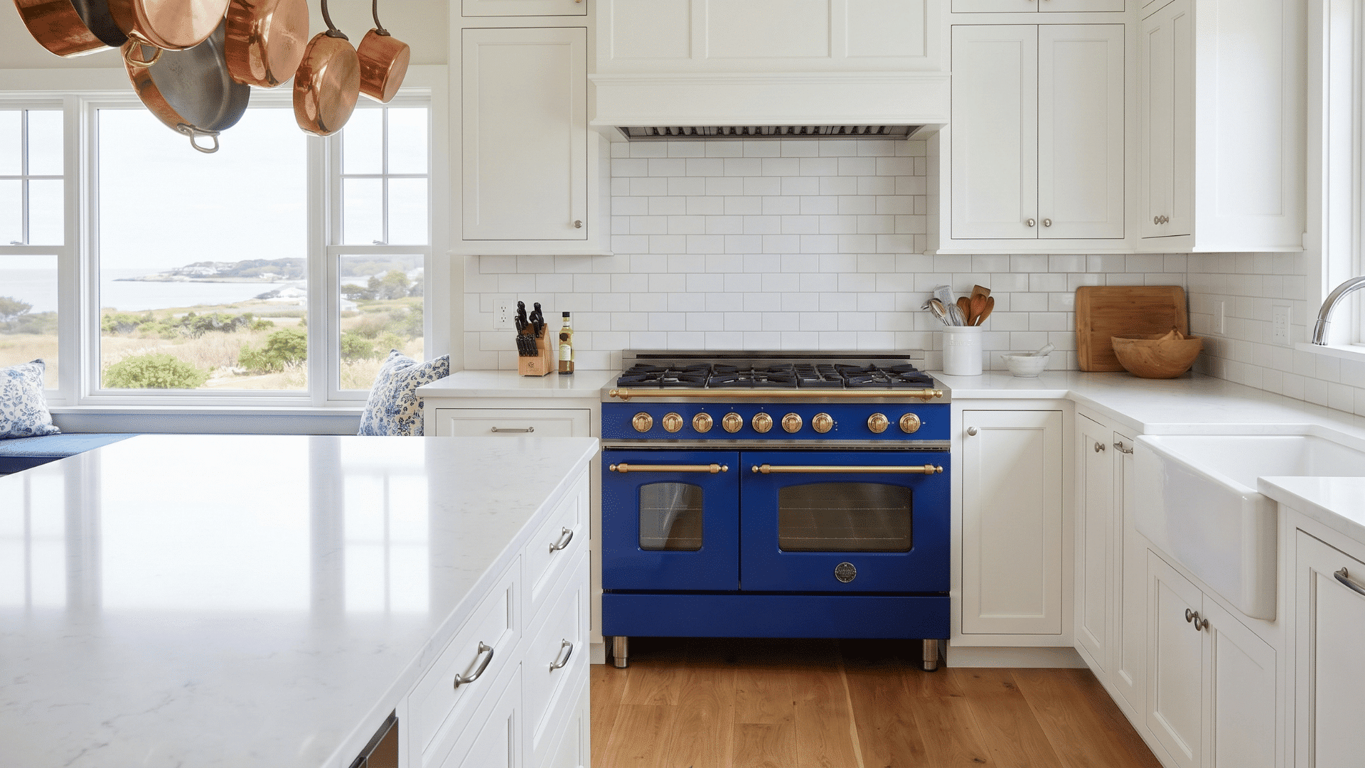 White upper and navy lower cabinets with a blue and gold stove, white countertops, and copper pots hanging in a bright kitchen.