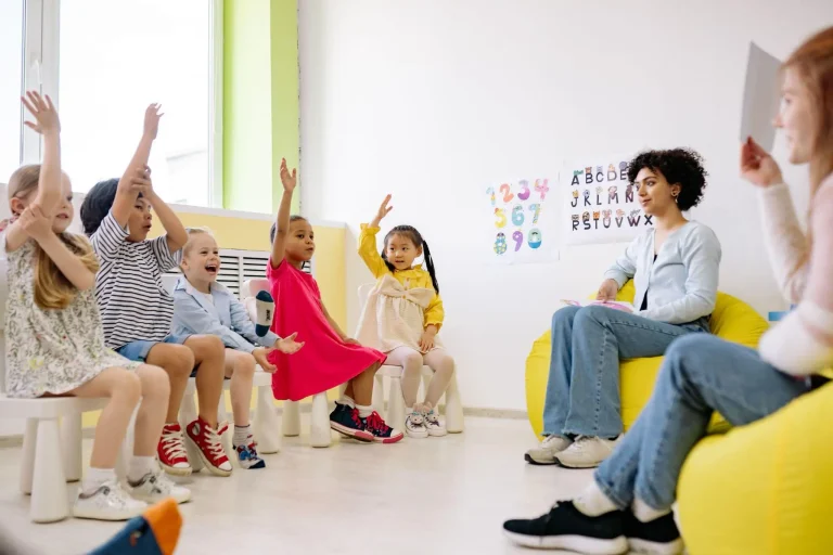 Children raising hands in a classroom with teacher sitting on a bean bag chair