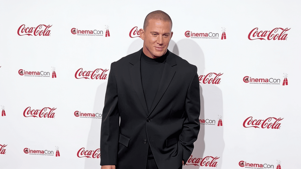 Actor Channing Tatum in a black suit jacket and turtleneck, posing against a white CinemaCon and Coca-Cola backdrop.