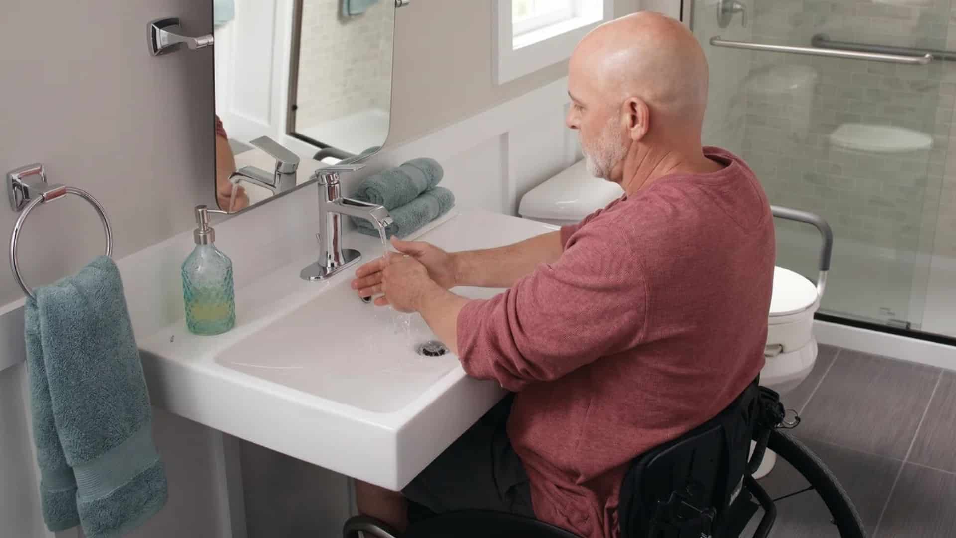 A man in a wheelchair washes his hands at an ADA-compliant wall-mounted bathroom sink with knee clearance.