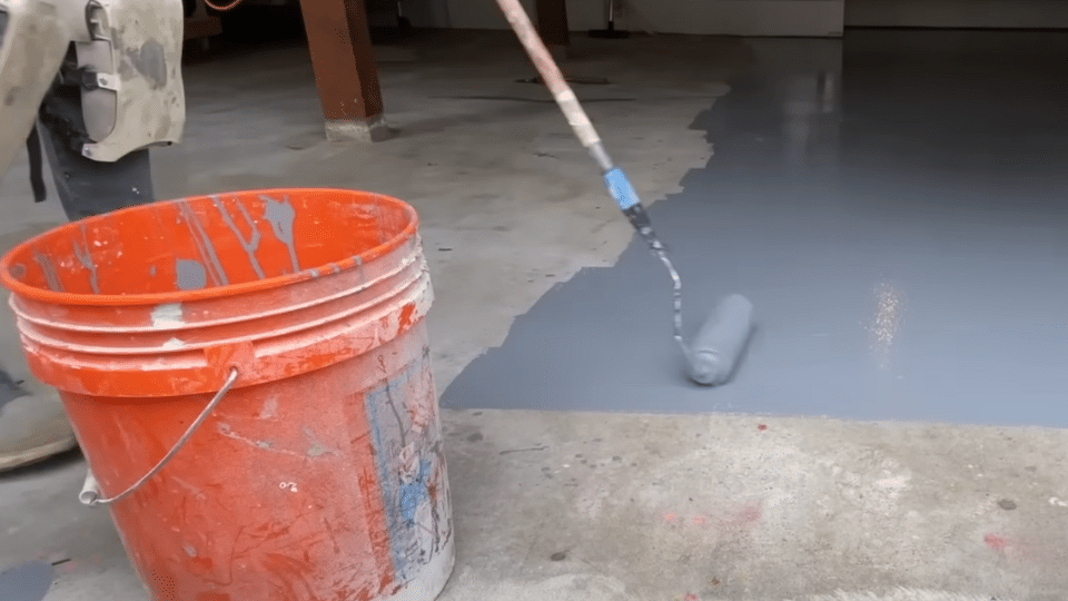 A person using a paint roller attached to an extension pole to apply grey epoxy coating on a garage floor next to an orange bucket.