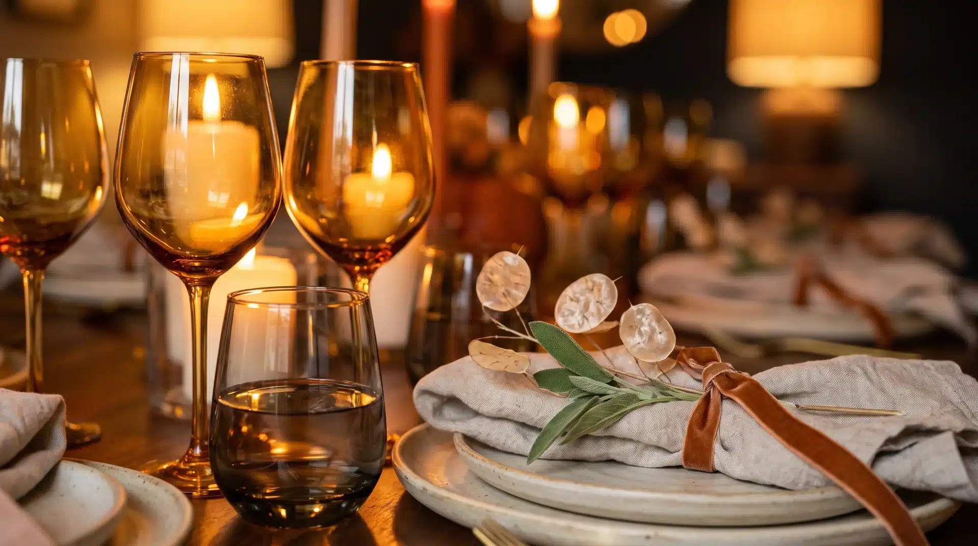 Amber wine glasses and smoked gray water glass glowing in candlelight beside a linen napkin with copper ribbon and sage.