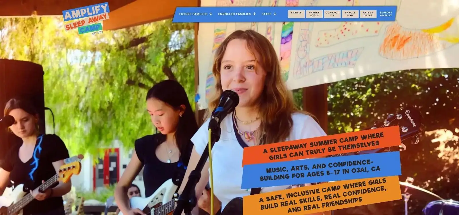 Three young girls playing guitars and singing into microphones outdoors under colorful banners
